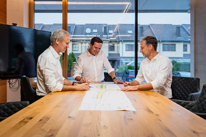 henninger-geschaeftsleitung-team Drei Männer diskutieren gemeinsam über Pläne auf einem großen Holztisch in einem lichtdurchfluteten Raum